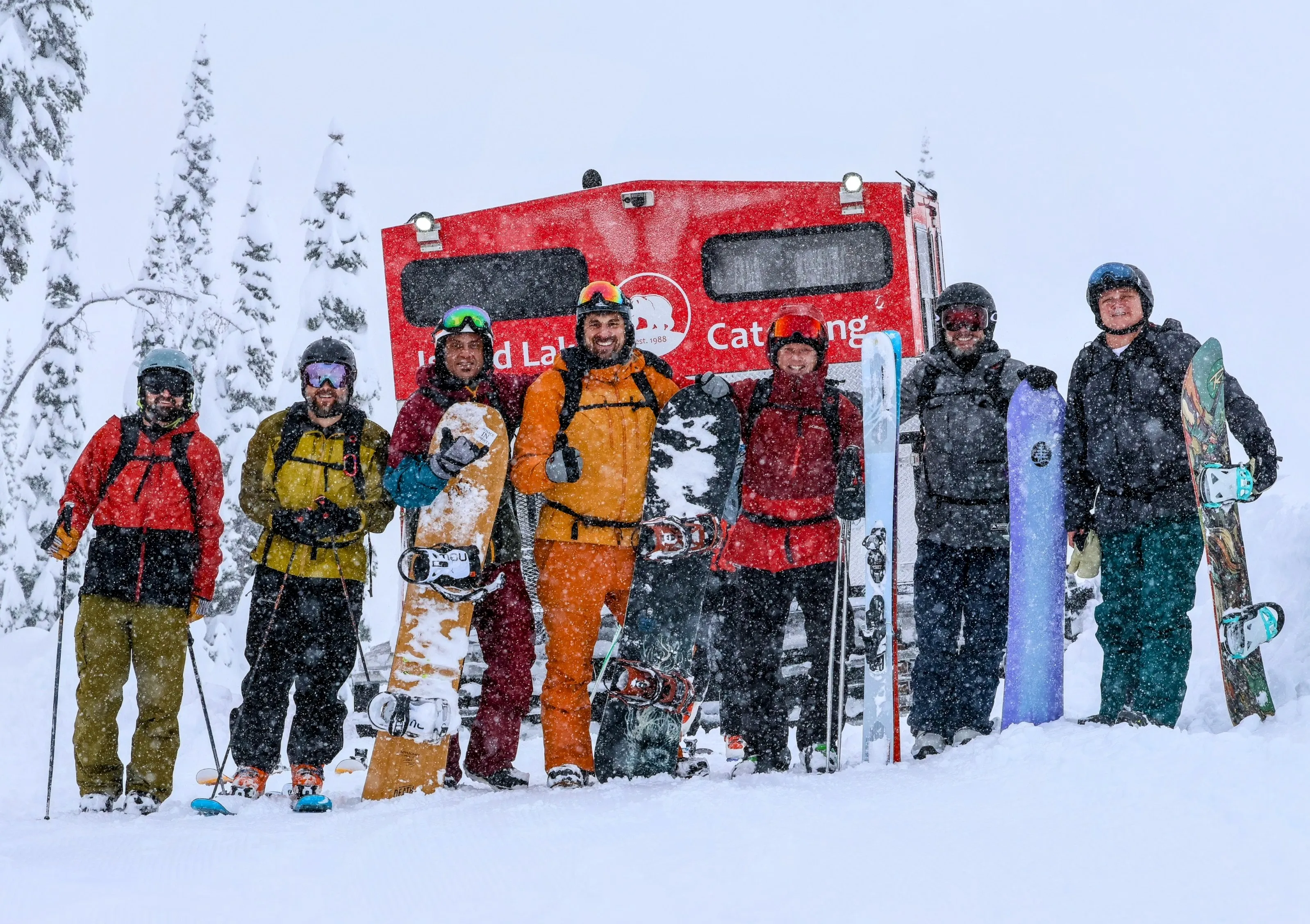 8 skiers and snowboarders lined up in falling snow beside a red catskiing snowcat.