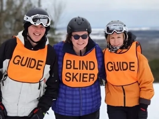 3 skiers in orange bibs, the middle one reading BLIND SKIER and the 2 either side reading GUIDE.